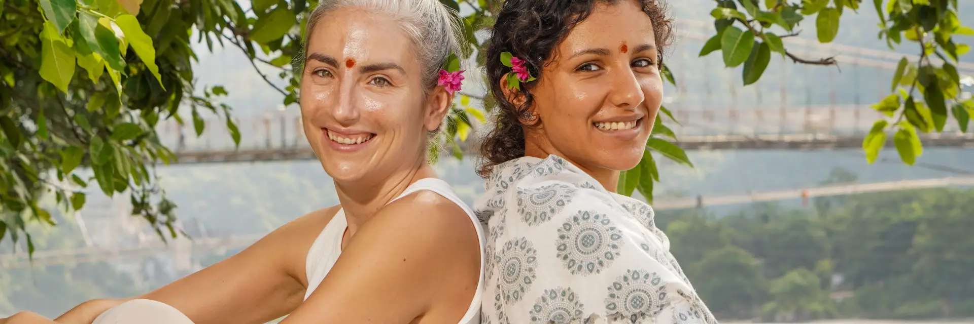 Two women sitting back-to-back, smiling, wearing white clothing and flower accents in their hair, with a blurred background of a river and bridge.